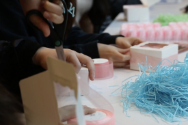 Person holding scissors to cut pink ribbon. Another person wrapping small white box with ribbon, blue straw paper in background. Person holding scissors to cut pink ribbon. Another person wrapping small white box with ribbon, blue straw paper in background.